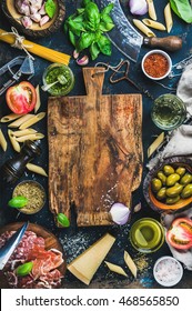 Italian Food Cooking Ingredients On Dark Background With Rustic Wooden Chopping Board In Center, Top View, Copy Space, Vertical Composition