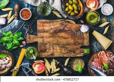 Italian Food Cooking Ingredients On Dark Background With Rustic Wooden Chopping Board In Center, Top View, Copy Space