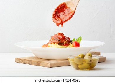 Italian Pasta In A White Plate. Pour Tomato Sauce With A Wooden Spoon. Olives, Basil Leaves. Stands On A Wooden Board. White Concrete Background. Horizontal. Direct View.