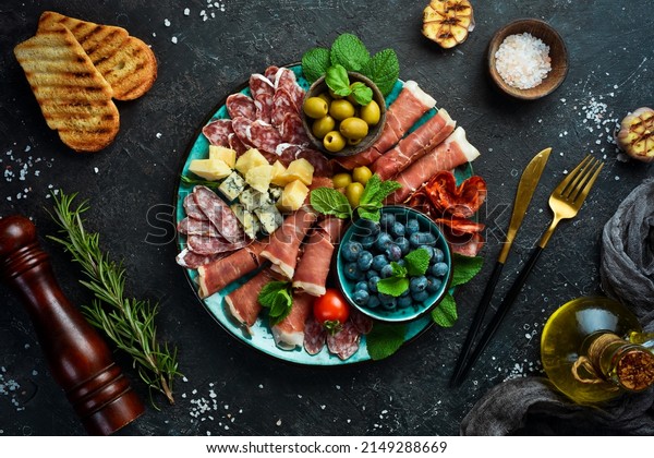 Italian snacks.\
Plate with cheese and ham, prosciutto, jamon salami, and snacks. On\
a black stone\
background.