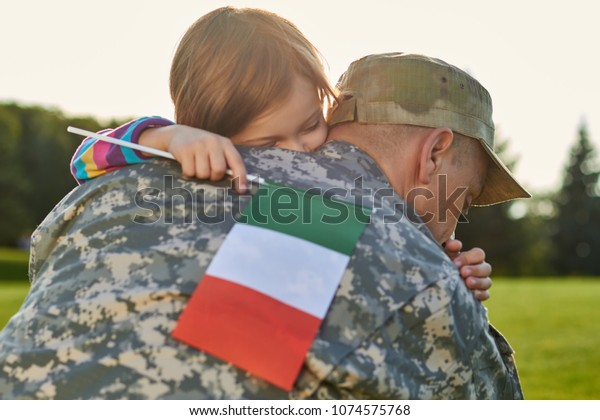 Italian soldier is hugging his little daughter.\
Little caucasian girl with italian flag is embracing her daddy in\
military uniform.