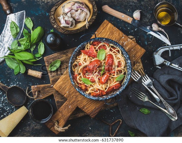 Italian style pasta dinner.\
Spaghetti with tomato and basil in plate on wooden board and\
ingredients for cooking pasta over dark plywood background, top\
view