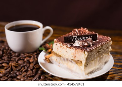 Italian stylish dessert with a piece of Tiramisu cake on a white saucer and a cup of black coffee surrounded by roasted beans scattered on a wooden table