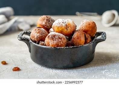 Italian traditional carnival fritters made with raisins and topped with sugar powder set in black pot on concrete table, strainer, napkin, raisins. Dark background. Close up - Shutterstock ID 2256992377