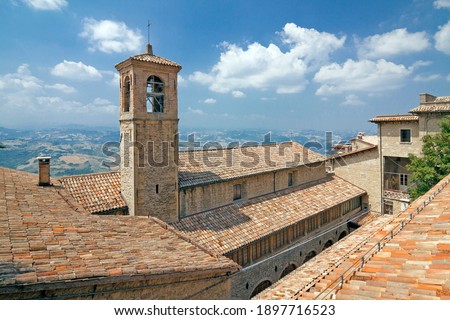 Italy - San Marino - The view of San Francisco church (1351) belfry with cross on roof against a majestic hilly landscape and dramatic tender cloudy blue sky background
