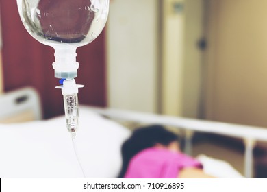 IV Drip Chamber, Tubing, And Bag Of Solution Against Blur Asian Patient Girl Resting On Hospital Bed.Inpatient During Treatment Concept.In Selective Focus.Blue Vintage Tone