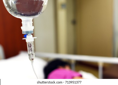 IV Drip Chamber, Tubing, And Bag Of Solution Against Blur Asian Patient Girl Resting On Hospital Bed.Inpatient During Treatment Concept.In Selective Focus.Blue Vintage Tone