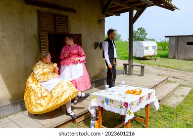 Ivanovo, Vojvodina, Serbia - April 17, 2016: Two Girls With One Guy Wearing A Traditional Folk Costume, Are Enjoy Eating Fresh Baked Donuts With Sugar At Outdoor In Shade.