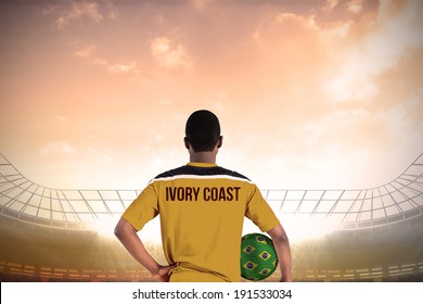 Ivory Coast Football Player Holding Ball Against Large Football Stadium Under Cloudy Blue Sky