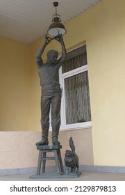 Izhevsk, Russia - July 12, 2018: Bronze Sculpture Of A Working Electrician Who, Standing On A Stool, Screws A Light Bulb Into A Ceiling Lamp. There Is A Cat On The Floor Looking At It.             