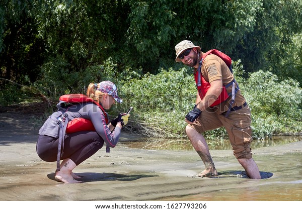 IZMAIL, UKRAINE - July 13, 2019: Man\
and woman standing in natural quicksand river, clay sediments,\
sinking, drowning quick sand, stuck in the\
soil