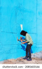 Izmir, Turkey, October 27, 2021: Black-haired Woman Painting A Wall Paints The Blue Building In Front Of The Ancient City Of Agora In Çankaya District. Unidentified Mural Painter. Remote Shooting.