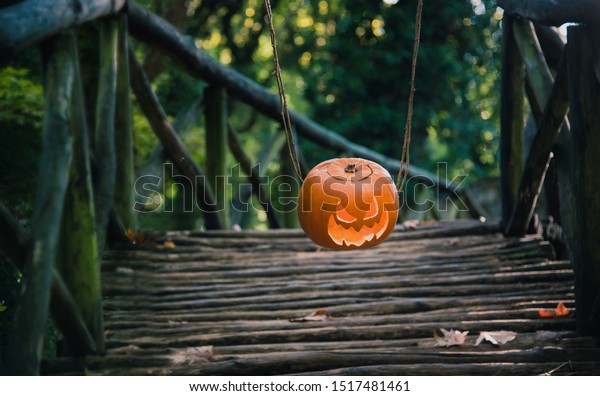Jack o\
lantern with creepy face hanging on lianas in the forest above\
wooden bridge. Halloween carved pumpkin head.\
