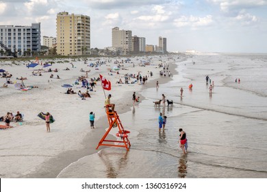 JACKSONVILLE BEACH, FL, USA/MARCH 15, 2019: A Young Lifeguard On Duty At Water's Edge Keeps Watch On Waders And Swimmers Within Sight Of The American Red Cross Flag Above His Orange Lifeguard Chair.