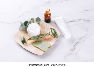 Jade facial roller and gua sha tool with beauty serum, face oil and face moisturizer on white marble table background. Facial massage kit for lifting massage therapy, home spa, mock up