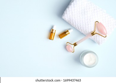 Jade roller for face massage with essential oil bottles, towel, cream moisturizer on blue background. Flat lay, top view, copy space. Rose quartz facial massage tool, beauty tool for skin care concept