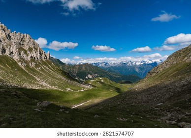 Jagged Alpine Cliffs Frame a Sunlit Mountain Basin Beneath Brilliant Summer Skies - A dramatic alpine landscape unfolds between steep rocky slopes and sharp limestone cliffs overlooking a broad mountain basin. Sunlight breaks through scattered clouds and illuminates rolling green terrain below, creating a striking contrast between bright meadows and shaded mountainsides. In the distance, a chain of snow-covered peaks stretches across the horizon, adding depth and scale to the panoramic scene. The combination of rugged rock formations, open grasslands, and high-altitude wilderness captures the grandeur of the mountain environment during a clear summer day.