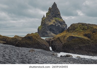 A jagged sea stack rises from the ocean, partially covered in moss, with a black pebble beach and rugged rocks under an overcast sky in Iceland.