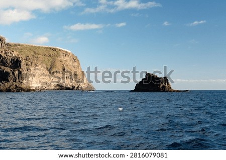A jagged volcanic sea stack stands off the layered cliffs topped by a small clifftop settlement on the deep blue Atlantic coast of La Gomera under a bright sky.