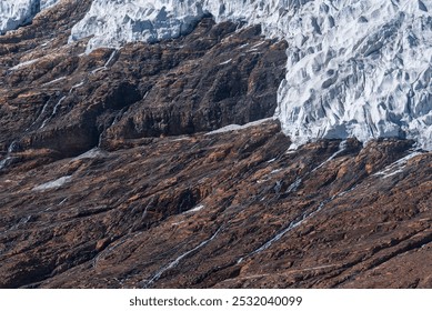 Jagged, white glacier cascades down rugged, dark brown rock face, creating a striking contrast between ice and earth. - Powered by Shutterstock