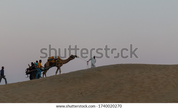 JAISALMER, RAJASTHAN / INDIA,
NOVEMBER 2018 : A cameleer (Camel Driver) carrying the decorated
Camel Cart on sam sand dunes of Thar desert with tourists during
sunset