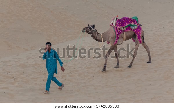 JAISALMER, RAJASTHAN / INDIA,\
NOVEMBER 2018 : A cameleer (Camel Driver) carrying the decorated\
Camel Cart on sam sand dunes of Thar desert with tourists during\
sunset