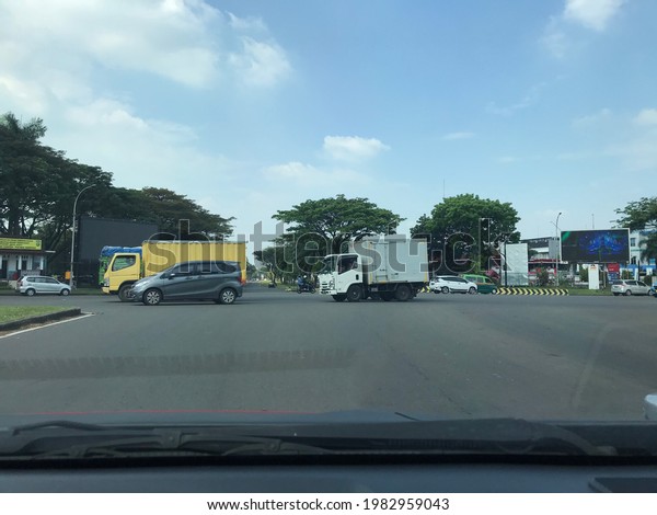 Jakarta 19 May 2021 - Intersection highway\
with many vehicles such as cars and trucks passing by, as well as\
tall trees on the side of the road and a giant outdoor LED screen\
displaying\
advertisements