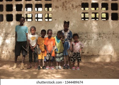 Jali, Gambia, Africa, May 18, 2017:  Horizontal Photo Of A Group Of Young School Kids In Colorful Clothes And New Shoes Standing In Front Of An Old Wall, Outdoors On A Sunny Day, Looking Into Camera