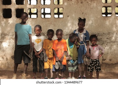 Jali, Gambia, Africa, May 18, 2017:  Horizontal Photo Of A Group Of Young School Kids In Colorful Clothes And New Shoes Standing In Front Of An Old Wall, Outdoors On A Sunny Day, Looking Into Camera