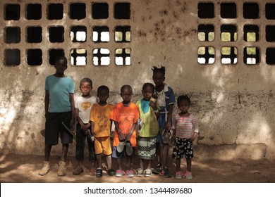 Jali, Gambia, Africa, May 18, 2017:  Horizontal Photo Of A Group Of Young School Kids In Colorful Clothes And New Shoes Standing In Front Of An Old Wall, Outdoors On A Sunny Day, Looking Into Camera