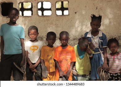 Jali, Gambia, Africa, May 18, 2017:  Horizontal Photo Of A Group Of Young School Kids In Colorful Clothes And New Shoes Standing In Front Of An Old Wall, Outdoors On A Sunny Day, Looking Into Camera