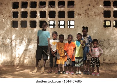 Jali, Gambia, Africa, May 18, 2017:  Horizontal Photo Of A Group Of Young School Kids In Colorful Clothes And New Shoes Standing In Front Of An Old Wall, Outdoors On A Sunny Day, Looking Into Camera