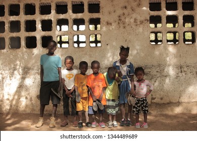 Jali, Gambia, Africa, May 18, 2017:  Horizontal Photo Of A Group Of Young School Kids In Colorful Clothes And New Shoes Standing In Front Of An Old Wall, Outdoors On A Sunny Day, Looking Into Camera