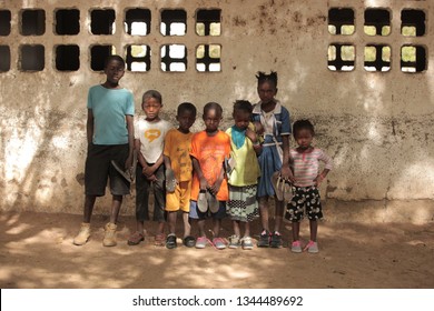 Jali, Gambia, Africa, May 18, 2017:  Horizontal Photo Of A Group Of Young School Kids In Colorful Clothes And New Shoes Standing In Front Of An Old Wall, Outdoors On A Sunny Day, Looking Into Camera