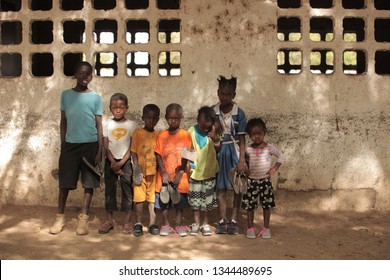 Jali, Gambia, Africa, May 18, 2017:  Horizontal Photo Of A Group Of Young School Kids In Colorful Clothes And New Shoes Standing In Front Of An Old Wall, Outdoors On A Sunny Day, Looking Into Camera