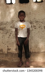 Jali, Gambia, Africa, May 19, 2017:   Vertical  Photography Of A Young Boy In Colorful Clothes And New Shoes Standing In Front Of School Wall, Outdoors On A Sunny Day, On Sandy Ground