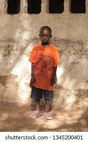 Jali, Gambia, Africa, May 19, 2017:   Vertical  Photography Of A Young Boy In Colorful Clothes And New Shoes Standing In Front Of School Wall, Outdoors On A Sunny Day, On Sandy Ground