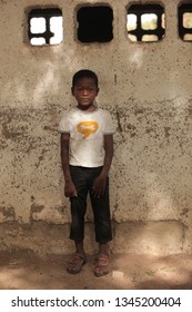 Jali, Gambia, Africa, May 19, 2017:   Vertical  Photography Of A Young Boy In Colorful Clothes And New Shoes Standing In Front Of School Wall, Outdoors On A Sunny Day, On Sandy Ground