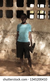 Jali, Gambia, Africa, May 19, 2017:   Vertical  Photography Of A Young Boy In Colorful Clothes And New Shoes Standing In Front Of School Wall, Outdoors On A Sunny Day, On Sandy Ground