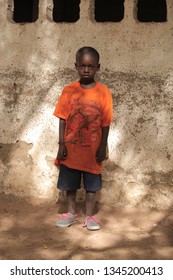 Jali, Gambia, Africa, May 19, 2017:   Vertical  Photography Of A Young Boy In Colorful Clothes And New Shoes Standing In Front Of School Wall, Outdoors On A Sunny Day, On Sandy Ground