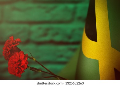 Jamaica Flag With Two Red Carnation Flowers For Honour Of Veterans Or Memorial Day On Teal, Sea-green Blurred Painted Brick Wall. Jamaica Glory To The Heroes Of War Concept.