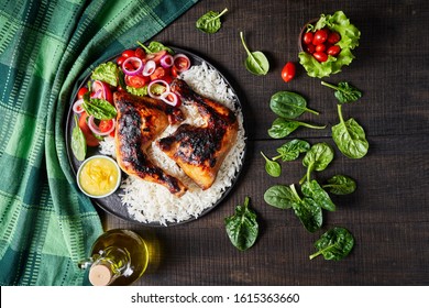 Jamaican chicken quarters chargrilled served on boiled rice with vegetables: tomatoes, lettuce, red onion, and spinach with orange sauce on a blue plate on rustic wooden background, top view 