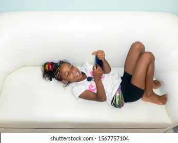 Jamaican European Ethnicity Little Girl With Colored Braids Lying On The Sofa Is Playing With The Mobile