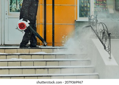 A Janitor Cleans Snow From The Stairs Next To The Shop With A Wind Blower. Lumps Of Snow And Snowflakes In The Air. Snow Cloud. Work Outdoors In Winter. Snow Removal After A Snowfall. Selective Focus.