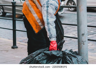 A Janitor In Red Gloves And Yellow Uniform On The City’s Street Takes A Few Big Black Bags Of Trash. Cleaner. Job. Working. Urban. Outdoor