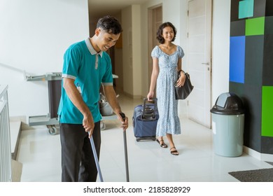 Janitor Wearing Turquoise Uniform Sweeps As Female Guests Walk Past In Hotel Lobby