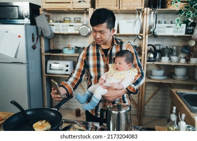 Japanese Asian New Dad Holding A Smartphone Is Taking A Photo With His Cute Baby Girl In Arm While Cooking Breakfast In The Kitchen At Home