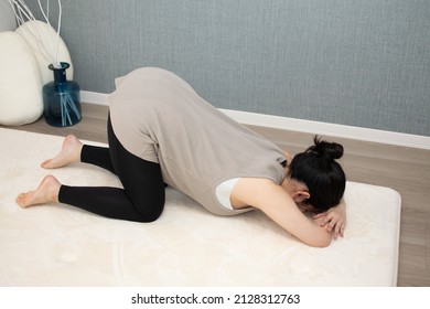 A Japanese Asian young beautiful pregnant woman lying on her stomach with relaxed clothes exercises regime to convert breech presentation on a white carpet with blue background from side angle