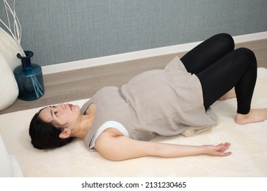 A Japanese Asian young beautiful pregnant woman lying on her back with relaxed clothes exercises regime to convert breech presentation on a white carpet with blue background from side angle