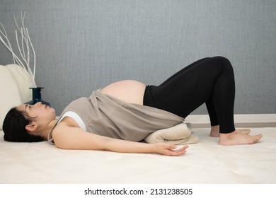 A Japanese Asian young beautiful pregnant woman lying on her back with relaxed clothes pulls up her clothes exercises regime to convert breech presentation on a white carpet with blue background from 